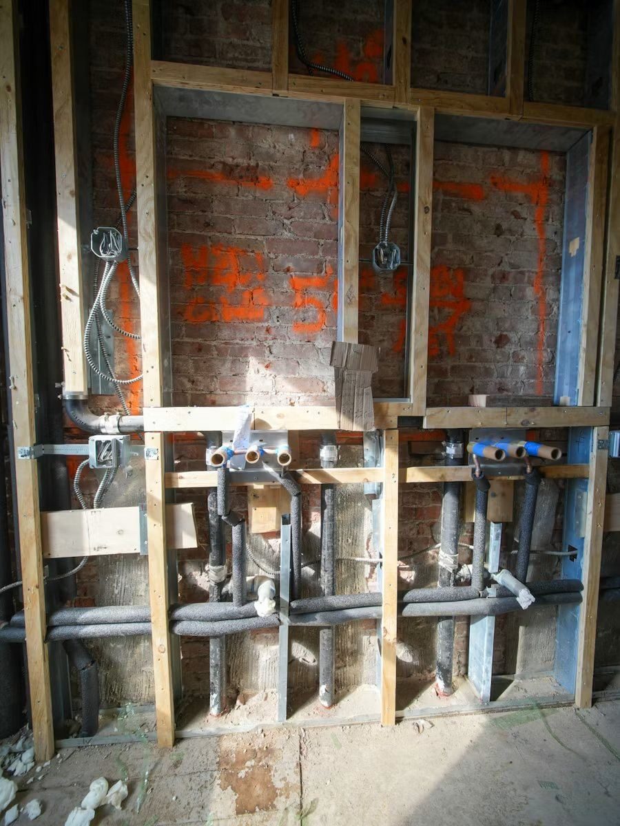 In-wall plumbing rough-in showing PVC pipes, fittings, and flexible conduit against exposed brick during construction